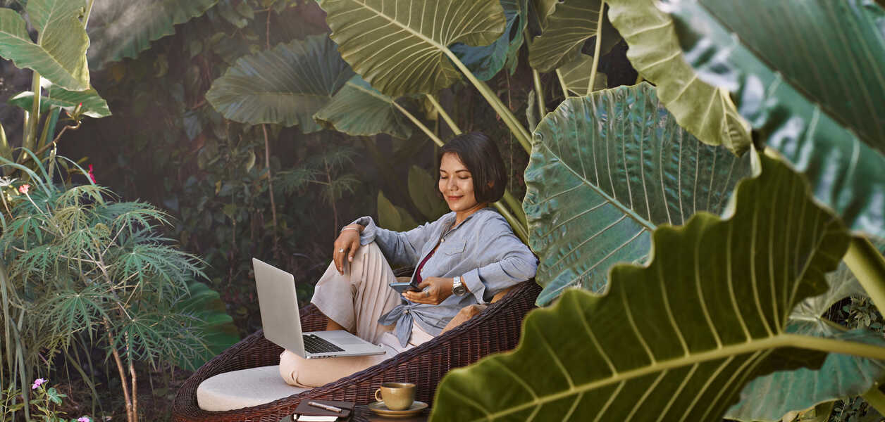 Asian woman working from home sitting in garden furniture surrounded by tropical plants, using laptop computer and mobile phone