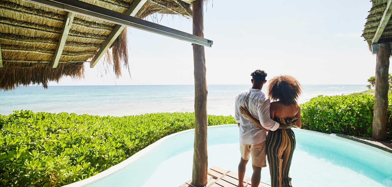 Wide shot rear view of couple standing poolside at luxury tropical beachfront villa looking at view